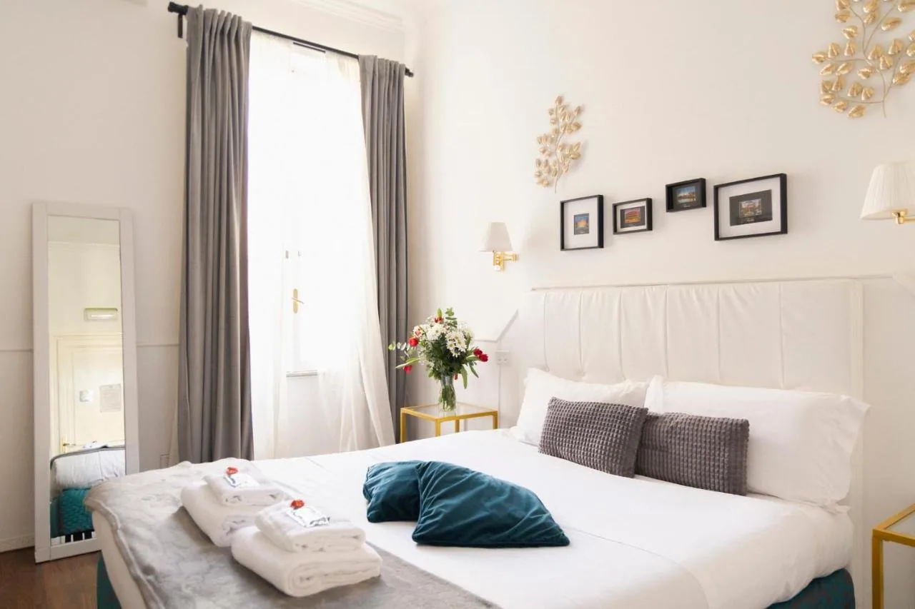Camera di Suitedream Vatican con letto matrimoniale bianco, tende grigie, fiori freschi e dettagli dorati